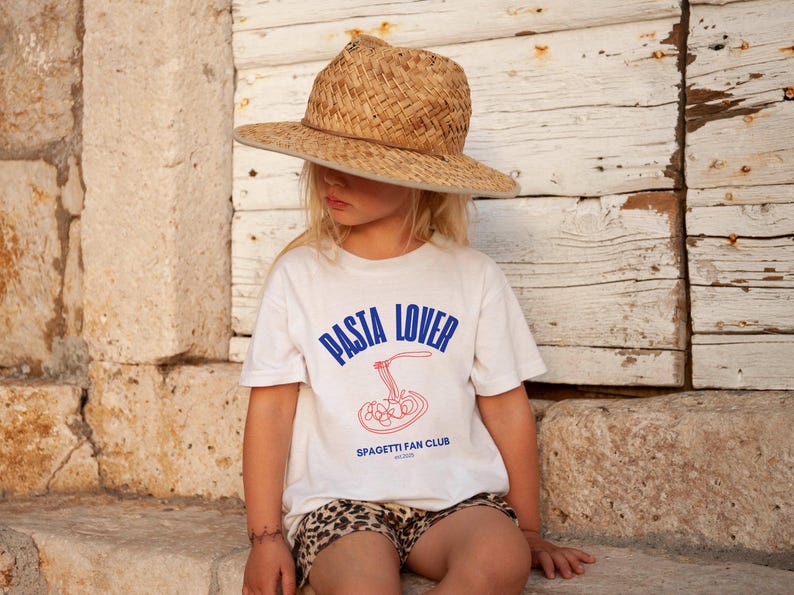 May include: A young person wearing a white t-shirt with the text "PASTA LOVER" and "SPAGHETTI FAN CLUB" in blue. The person is wearing a straw hat and has a brown and white animal print shorts on.