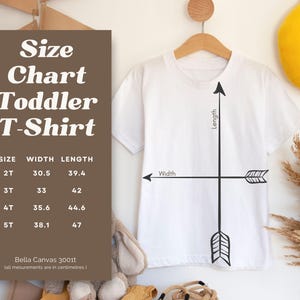 May include: Size chart for toddler t-shirts with measurements in inches. The chart shows the width and length of the shirts in sizes 2T, 3T, 4T, and 5T. The chart is printed on a brown background with white text. A white t-shirt with a black arrow graphic is shown in the image.