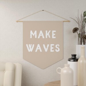 May include: A beige fabric banner with the text "MAKE WAVES" in white paint. The banner is hanging from a string and is displayed against a white wall.