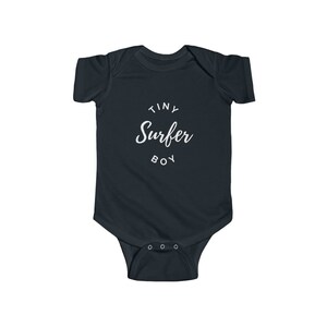 Puede incluir: Un body de beb&eacute; azul oscuro con texto blanco que dice "Tiny Surfer Boy" en un c&iacute;rculo.