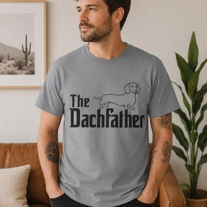 Op de afbeelding: Een grijs t-shirt met korte mouwen voorzien van een zwarte afbeelding van een teckel en de tekst "The Dachfather" op de voorzijde. Het t-shirt heeft een ronde hals en een standaard pasvorm, gemaakt van een effen gekleurde stof.
