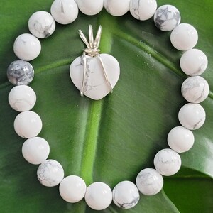 May include: A white beaded bracelet with a heart-shaped pendant. The pendant is wrapped in silver wire. The beads are round and white with gray marbling. The bracelet and pendant are on a green leaf.
