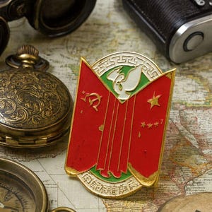 Puede incluir: Imagen de temática vintage con un pin rojo y dorado con un diseño de pájaro blanco, sobre un mapa. Otros elementos incluyen una brújula, unos prismáticos, un reloj de bolsillo y una cámara, sugiriendo un tema de viaje o exploración.