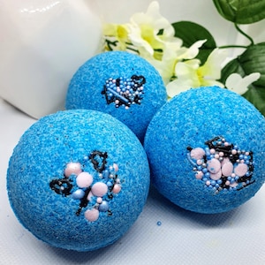 May include: Three spherical, blue bath bombs with a textured finish. Each bath bomb is adorned with a design featuring pink, blue, and black details. The bath bombs are displayed on a white surface, with flowers visible in the background.