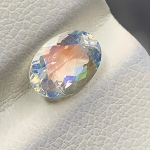Puede incluir: Una gema ovalada con iridiscencia de arco&iacute;ris. La gema muestra un espectro de colores, incluyendo rosa, azul y amarillo, y est&aacute; colocada sobre una superficie blanca y texturizada. La gema est&aacute; facetada, reflejando la luz.