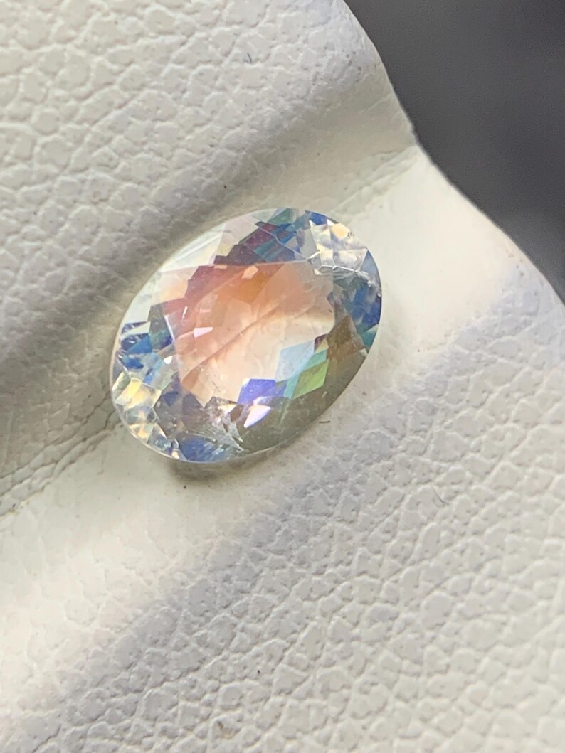 Puede incluir: Una gema ovalada con iridiscencia de arco&iacute;ris. La gema muestra un espectro de colores, incluyendo rosa, azul y amarillo, y est&aacute; colocada sobre una superficie blanca y texturizada. La gema est&aacute; facetada, reflejando la luz.