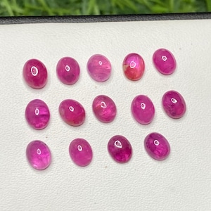 May include: A collection of oval-shaped, polished ruby gemstones. The rubies display a vibrant pinkish-red hue, with variations in color intensity. The gemstones are arranged on a white surface, with a blurred green background.