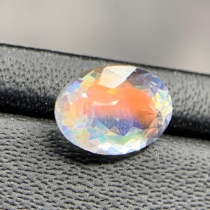 Puede incluir: Una gema ovalada con una superficie facetada. La gema muestra un espectro de colores, incluyendo naranja, azul y amarillo, con una apariencia clara e iridiscente. La gema est&aacute; sobre un fondo oscuro y texturizado.