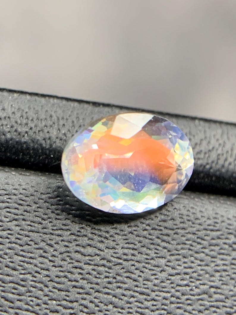 Puede incluir: Una gema ovalada con una superficie facetada. La gema muestra un espectro de colores, incluyendo naranja, azul y amarillo, con una apariencia clara e iridiscente. La gema est&aacute; sobre un fondo oscuro y texturizado.