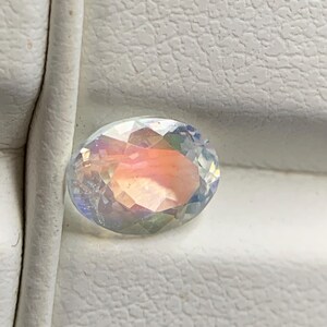 Puede incluir: Una gema ovalada e iridiscente con una superficie facetada. La gema muestra un espectro de colores, incluyendo rosa, naranja y azul, sobre un fondo blanco texturizado. La gema es probablemente un componente de joyer&iacute;a.