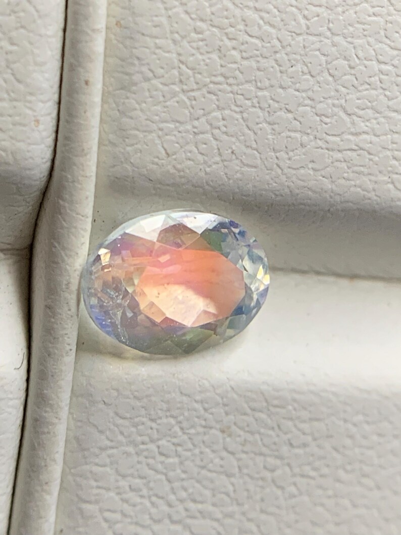Puede incluir: Una gema ovalada e iridiscente con una superficie facetada. La gema muestra un espectro de colores, incluyendo rosa, naranja y azul, sobre un fondo blanco texturizado. La gema es probablemente un componente de joyer&iacute;a.