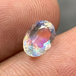 Puede incluir: Una gema de corte ovalado con iridiscencia de arco&iacute;ris. La superficie clara y facetada de la gema refleja la luz, mostrando un espectro de colores que incluye rosa, azul y amarillo. La gema se sostiene entre los dedos.