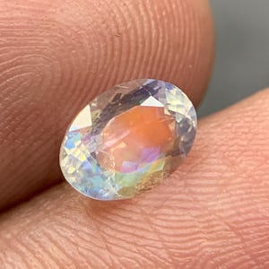 Puede incluir: Una gema de corte ovalado con iridiscencia de arco&iacute;ris. La superficie clara y facetada de la gema refleja la luz, mostrando un espectro de colores que incluye azul, amarillo y naranja. La gema se sostiene entre dos dedos.