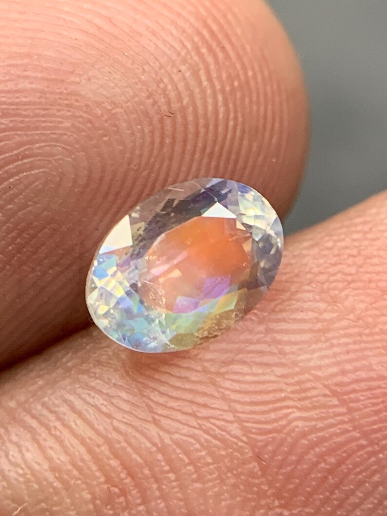 Puede incluir: Una gema de corte ovalado con iridiscencia de arco&iacute;ris. La superficie clara y facetada de la gema refleja la luz, mostrando un espectro de colores que incluye azul, amarillo y naranja. La gema se sostiene entre dos dedos.