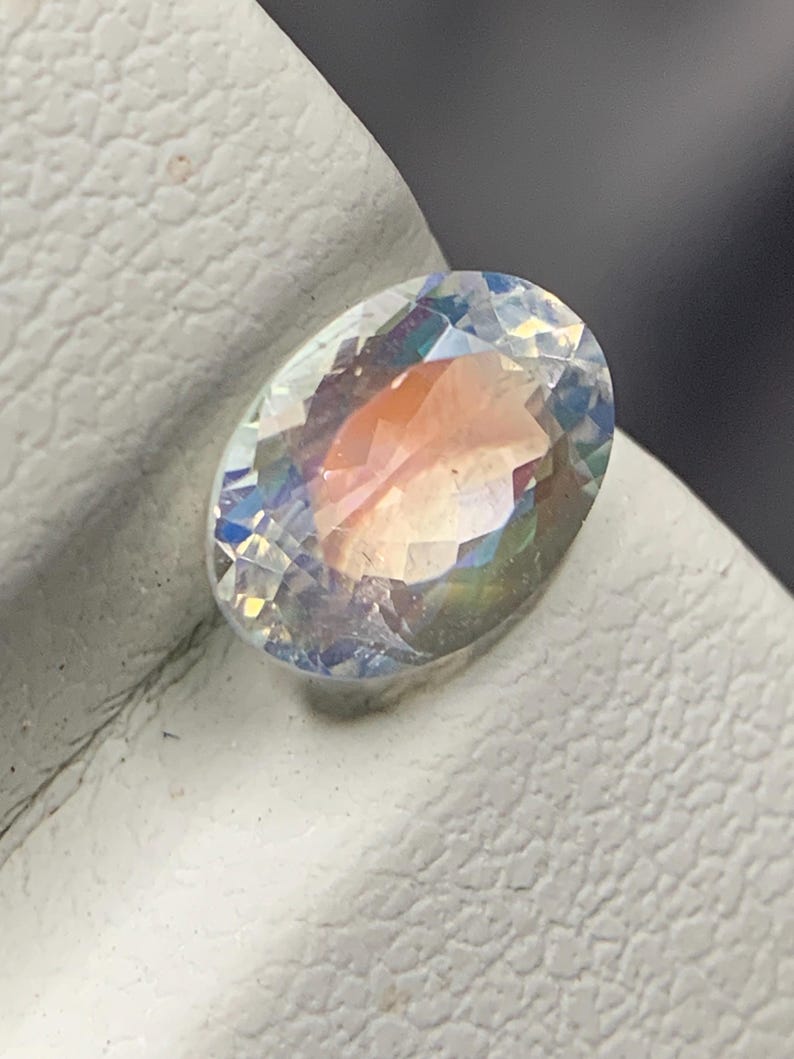 Puede incluir: Una gema de corte ovalado con un efecto iridiscente de arco&iacute;ris. La superficie facetada de la gema refleja la luz, mostrando un espectro de colores que incluye rosa, azul y amarillo. La gema se encuentra sobre un fondo blanco.