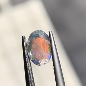 Puede incluir: Una gema de corte ovalado con iridiscencia de arco&iacute;ris, sujeta con unas pinzas plateadas. Las facetas de la gema reflejan tonos naranja, azul y verde, creando un juego de luces. El fondo est&aacute; borroso, enfocando la gema.
