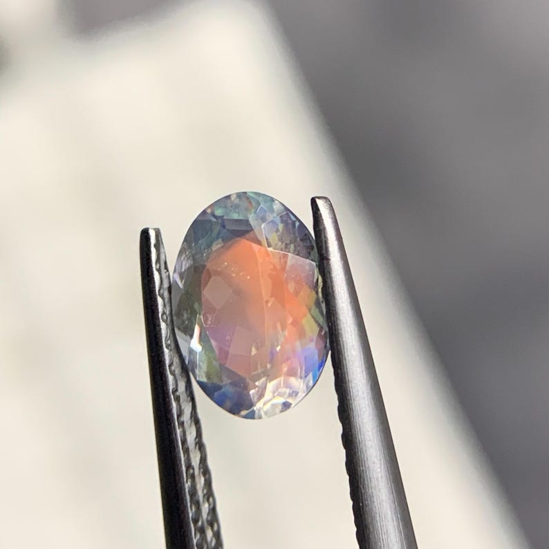 Puede incluir: Una gema de corte ovalado con iridiscencia de arco&iacute;ris, sujeta con unas pinzas plateadas. Las facetas de la gema reflejan tonos naranja, azul y verde, creando un juego de luces. El fondo est&aacute; borroso, enfocando la gema.