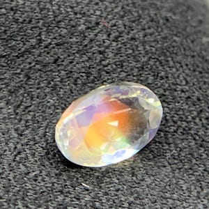 Puede incluir: Una gema ovalada con iridiscencia de arco&iacute;ris. La gema tiene una superficie clara y facetada que refleja un espectro de colores, incluyendo naranja, amarillo y morado. La piedra est&aacute; sobre un fondo oscuro y texturizado.