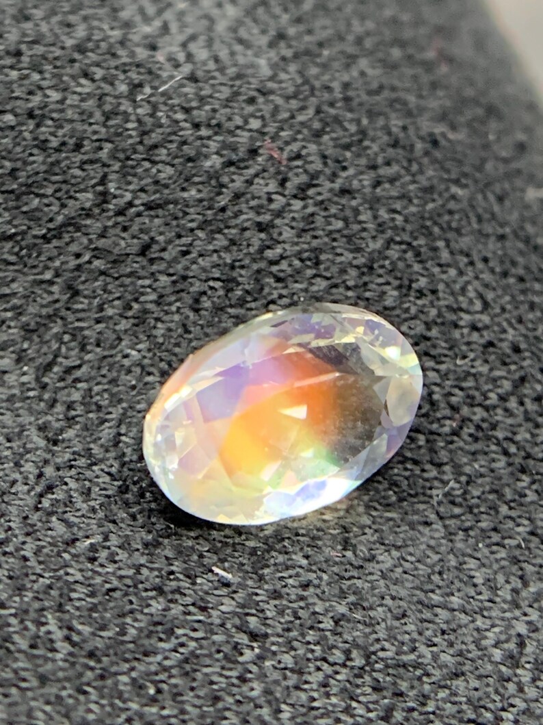 Puede incluir: Una gema ovalada con iridiscencia de arco&iacute;ris. La gema tiene una superficie clara y facetada que refleja un espectro de colores, incluyendo naranja, amarillo y morado. La piedra est&aacute; sobre un fondo oscuro y texturizado.