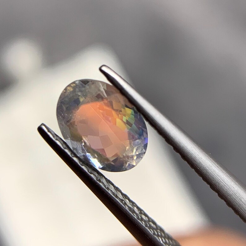 Puede incluir: Una gema de corte ovalado con iridiscencia de arco&iacute;ris, sujeta con unas pinzas. La gema tiene un interior melocot&oacute;n y amarillo con destellos azules y verdes. Las facetas de la piedra son claramente visibles.