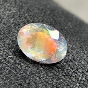 Puede incluir: Una gema de corte ovalado con iridiscencia de arco&iacute;ris. La superficie facetada de la gema captura y refleja la luz, mostrando un espectro de colores, incluyendo naranja, amarillo y verde. Sobre un fondo oscuro y texturizado.
