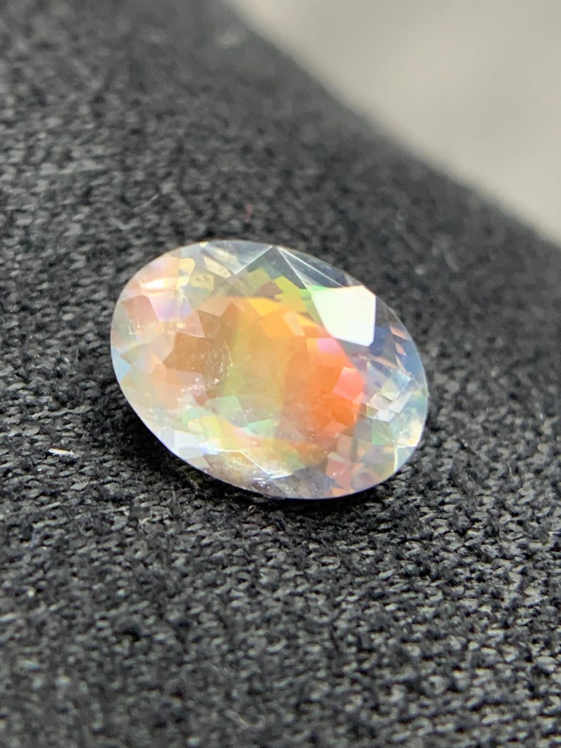 Puede incluir: Una gema de corte ovalado con iridiscencia de arco&iacute;ris. La superficie facetada de la gema captura y refleja la luz, mostrando un espectro de colores, incluyendo naranja, amarillo y verde. Sobre un fondo oscuro y texturizado.