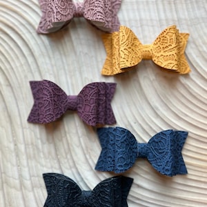 Könnte beinhalten: Fünf Haarschleifen mit Spitzenmuster in verschiedenen Farben: Mauve, Senfgelb, Dunkelviolett, Marineblau und Schwarz. Die Schleifen sind auf einer hellen Holzmaserung angeordnet, die ihre komplizierten Muster und Texturen hervorhebt. Elegantes Accessoire.