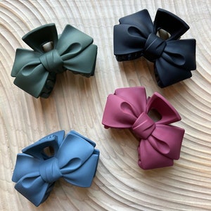 May include: Four bow-shaped hair clips in green, black, dusty rose, and blue. The clips are made of a matte material and are arranged on a light wood-grain surface. The bows have a classic, elegant design.