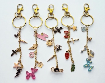 Può includere: Cinque portachiavi color oro con vari ciondoli. I ciondoli includono una croce, una stella, un fulmine, un cuore, ali d'angelo, un cane rosa, un ragno, un pipistrello, un fiocco, una fragola e un animale a palloncino. Ogni portachiavi ha un anello e un fermaglio dorati.