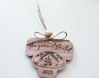 Può includere: Ornamento in legno con la scritta "The year it started" e l'anno 2025. L'ornamento presenta un design decorativo e un cordino per appenderlo, con una perlina di legno attaccata.