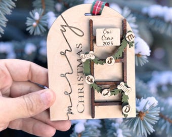 Può includere: Ornamento natalizio in legno con un design a scala. L'ornamento presenta le parole "Merry Christmas" e "Our Crew 2025". I nomi sono visualizzati sulla scala. Un nastro a quadri rosso e verde è attaccato per appenderlo.