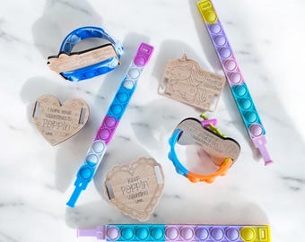 Può includere: Assortimento di braccialetti pop-it colorati con etichette di legno a forma di cuore. Le etichette hanno diversi messaggi di San Valentino, tra cui "Keep Poppin' Valentine!" e "You're Magical Valentine!"