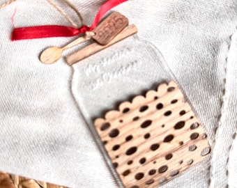 Può includere: Un ornamento trasparente a forma di barattolo con un design intagliato in legno all'interno. L'ornamento ha un cucchiaio di legno e un'etichetta con il numero "9885" legati con un nastro rosso e spago. Su un panno bianco.