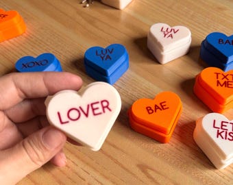 Può includere: Una collezione di scatole a forma di cuore in vari colori, tra cui bianco, blu e arancione. Ogni scatola ha un messaggio romantico diverso stampato, come "LOVER" e "BAE". Le scatole sono disposte su una superficie di legno.