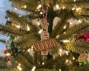 Può includere: Un ornamento per albero di Natale in legno con un motivo mimetico e il nome "Gunner" in corsivo. L'ornamento è appeso a un cordino a strisce rosse e bianche, sullo sfondo di un albero di Natale decorato.