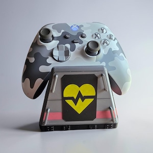 May include: A gray and white camouflage-patterned Xbox controller rests on a gray charging stand. The stand features a black panel with a yellow heart and a red line. The controller has a blue light.