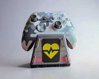 Può includere: Un controller Xbox grigio e bianco con motivo mimetico poggia su una base di ricarica grigia. Il supporto presenta un pannello nero con un cuore giallo e una linea rossa. Il controller ha una luce blu.
