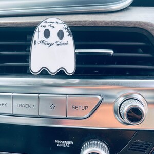 Op de afbeelding: Een witte geestenautoluchtverfrisser met de tekst "Always Tired" in zwarte letters. De luchtverfrisser hangt aan een autoventilatie.