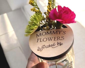 Può includere: Un cerchio di legno con il testo "Mommy's Flowers Picked by" è attaccato a un barattolo di vetro pieno di fiori gialli e verdi e un garofano rosa.