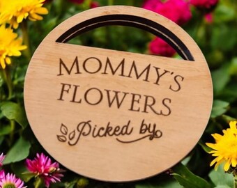 Può includere: Un cartello rotondo in legno con manico, inciso con le parole "MOMMY'S FLOWERS" e "Picked by" in un carattere corsivo. Il cartello è circondato da una vivace serie di fiori rosa, gialli e bianchi.