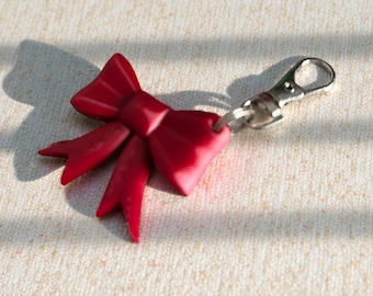 Può includere: Un portachiavi a forma di fiocco rosso con chiusura argentata. Il fiocco ha una finitura lucida e proietta un'ombra su una superficie strutturata bianco sporco. L'accessorio è decorativo.