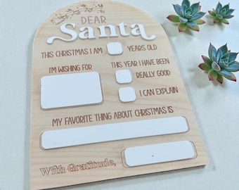 Può includere: Cartello in legno "Caro Babbo Natale" con testo bianco e sezioni da compilare. Il cartello include spazi per l'età, i desideri e le cose preferite del Natale. Sullo sfondo ci sono delle succulente decorative.