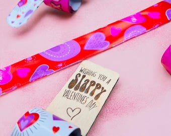 Può includere: Braccialetti schiaffo di San Valentino in rosso, rosa e bianco, decorati con cuori. Un braccialetto ha il testo "XOXO". Un'etichetta di legno recita "Wishing You a Slappy Valentine's Day" con un simbolo di cuore.