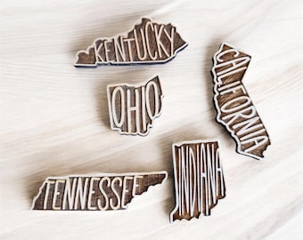 Può includere: Magneti in legno a forma di stati americani, tra cui Kentucky, Ohio, California, Tennessee e Indiana. Ogni magnete ha il nome dello stato ritagliato, creando un effetto a strati. I magneti sono disposti su una superficie di legno chiaro.