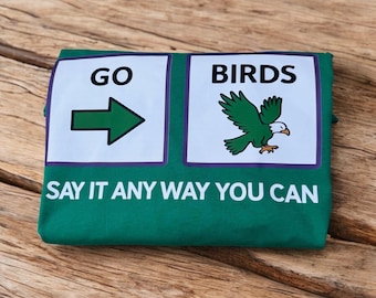 Può includere: T-shirt verde con le parole "GO" e una freccia, e "BIRDS" con una grafica di aquila. Il testo "SAY IT ANY WAY YOU CAN" è stampato sotto. La maglietta è piegata su una superficie di legno.