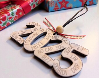 Può includere: Ornamento natalizio in legno a forma di 2025, inciso con vari termini gergali. L'ornamento ha un cordino nero e una piccola perla di legno per appenderlo. Regali confezionati sullo sfondo.