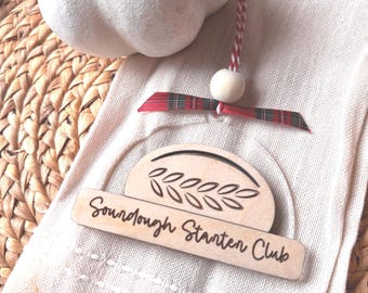 Può includere: Una zucca bianca con un accento di foglia d'oro e un'etichetta decorativa in legno con la scritta "Sourdough Starter Club". L'etichetta è attaccata a un panno bianco con un nastro scozzese rosso e verde.