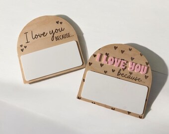 Può includere: Due targhe in legno con la scritta "I love you because..." in nero e rosa. Ogni targa ha uno spazio rettangolare bianco per scrivere e piccoli dettagli a forma di cuore. Le targhe sono su una superficie bianca.