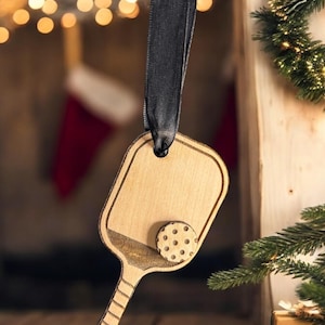 May include: A wooden pickleball paddle ornament with a black ribbon for hanging. The paddle is brown and has a white ball with black dots on it.