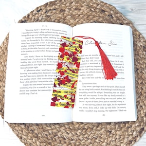 May include: A red and yellow crime scene do not cross bookmark with a red tassel. The bookmark is placed in an open book with the text "Chapter Six" visible.